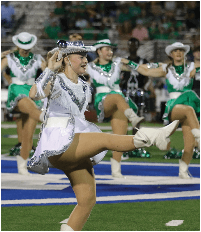 A group of people dressed in matching green and white dance uniforms perform a high kick routine on a sports field. Descripci n generada con IA