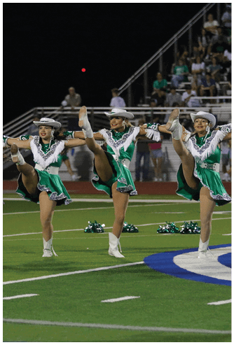 Three adults in green and white sequined dance costumes and white hats perform a high kick routine on a football field at night. Descripci n generada con IA