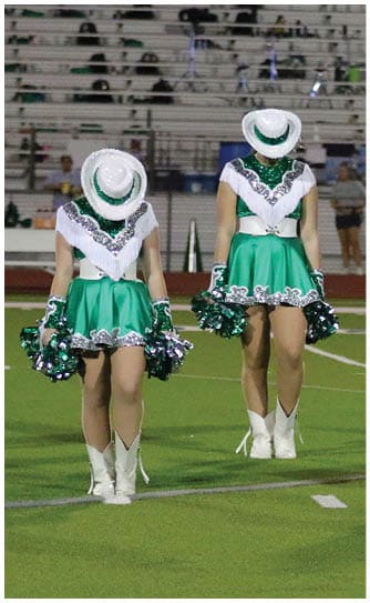 Adults in green and white sequined dance uniforms with white boots and hats stand on a football field holding green and silver pom-poms. Descripci n generada con IA