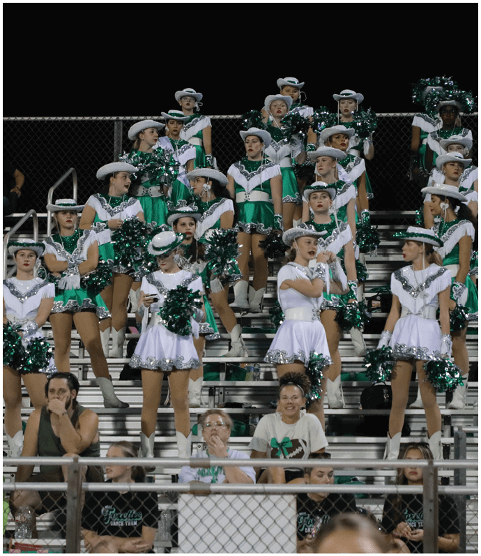A group of people dressed in green and white cheerleading or dance team uniforms with hats and pom-poms are standing and sitting on bleachers at an outdoor event at night. Descripci n generada con IA