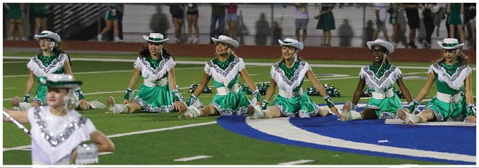 People dressed in green and white uniforms with hats are performing a synchronized routine on a sports field with spectators in the background. Descripci n generada con IA