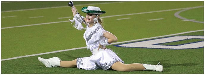 A person dressed in a white fringed outfit and a green and white hat is performing a dance routine on a football field. Descripci n generada con IA