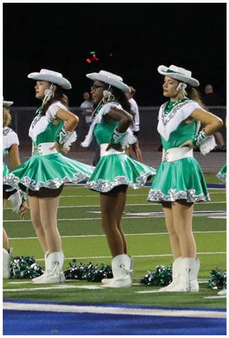 A line of people dressed in green and white cheerleading or dance uniforms with hats stand on a sports field at night. Descripci n generada con IA