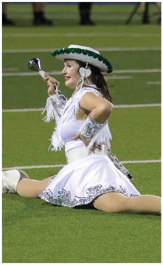A person dressed in a white fringed and decorated dance costume with a matching hat is kneeling on a sports field while holding a baton. Descripci n generada con IA
