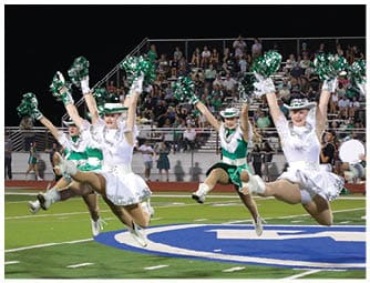 People dressed in white and green cheerleading uniforms are performing high jumps with pom-poms on a sports field in front of a seated crowd. Descripci n generada con IA