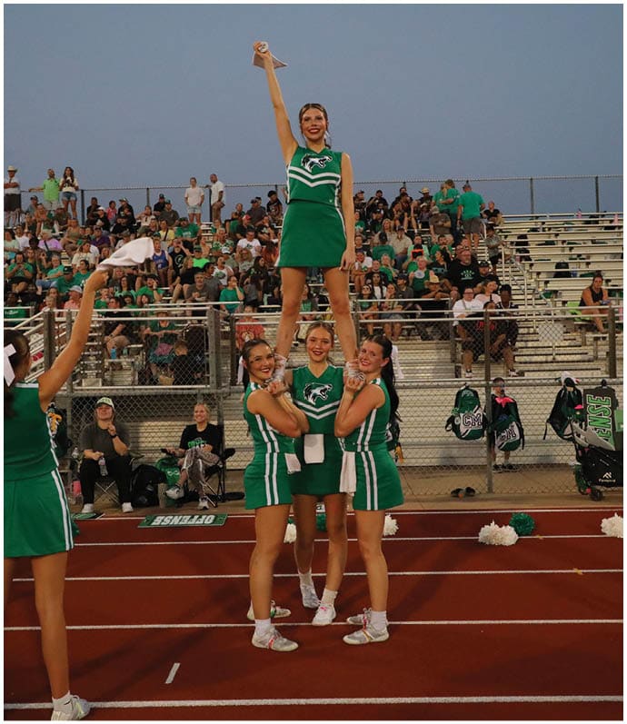 Four adults in green cheerleading uniforms perform a stunt on a track in front of bleachers filled with spectators. Descripci n generada con IA