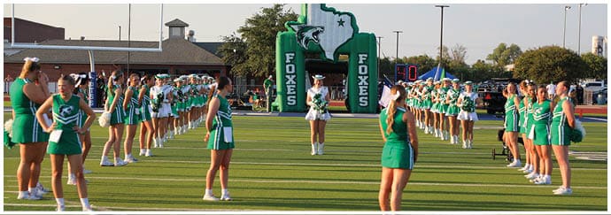 People in green and white cheerleading uniforms stand in two long lines facing each other on a sports field. Descripci n generada con IA