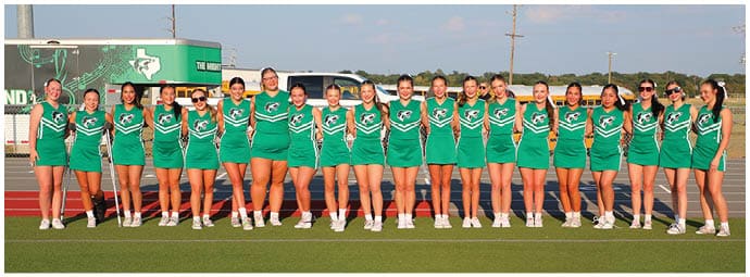 A group of people wearing matching green cheerleading uniforms stand in a line on a sports field with a scoreboard and vehicles in the background. Descripci n generada con IA