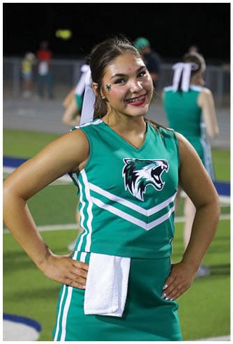 A person wearing a green cheerleading uniform with a wildcat logo and a white towel tucked into the waist is smiling on a sports field at night. Descripci n generada con IA