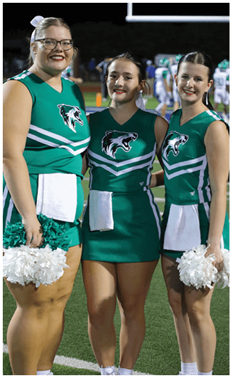 A person wearing a green cheerleading uniform with a wolf logo stands on a sports field with hands on hips and a white towel hanging from the waist. Descripci n generada con IA