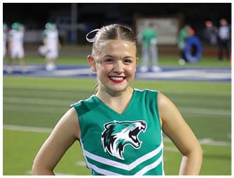 A person wearing a green cheerleading uniform with a white ribbon in their hair is smiling on a sports field at night. Descripci n generada con IA