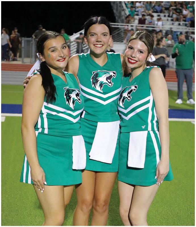Three people wearing green skirts, white socks, and white sneakers stand on a green sports field near a blue and white line. Descripci n generada con IA