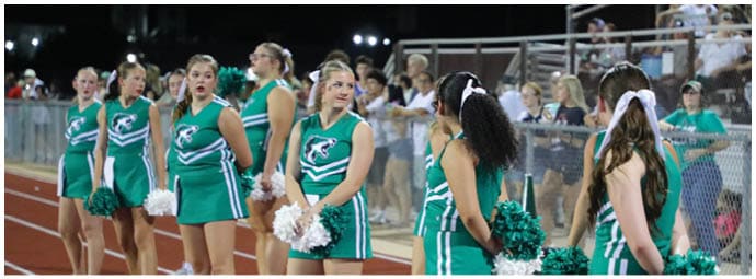 A line of people wearing green and white cheerleading uniforms stand on a track holding pom-poms. Descripci n generada con IA