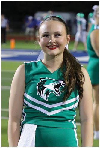 A person with long hair tied back is smiling and wearing a green cheerleading uniform with a fierce animal logo on the front, standing on a sports field at night. Descripci n generada con IA