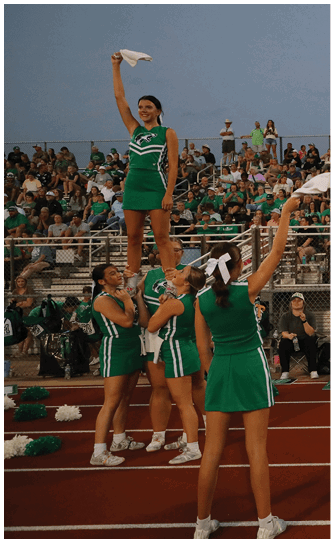 A group of cheerleaders in green uniforms perform on a track in front of a crowded stadium during an outdoor event. Descripci n generada con IA