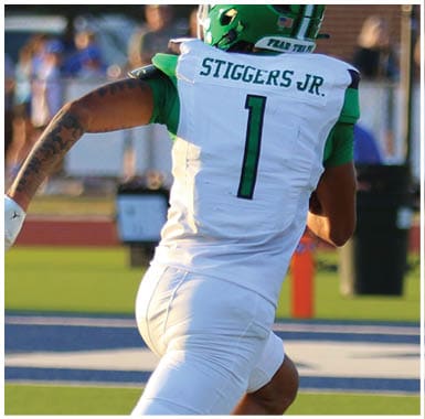 A person wearing a green helmet and white sports jersey with the name “STIGGERS JR." and the number 1 is preparing to throw a football on a field with blurred spectators in the background. Descripci n generada con IA