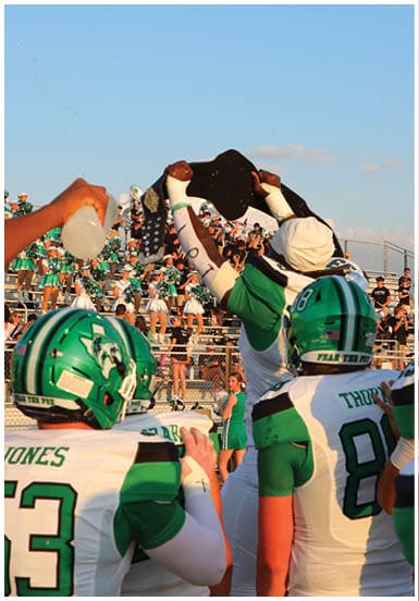 A group of people in green and white football uniforms celebrate on a field with one person holding a large black object above their head while spectators watch from bleachers in the background. Descripci n generada con IA