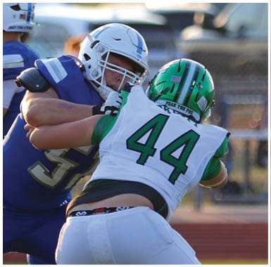 Two people in football gear are engaged in a physical play, with one wearing a white helmet and blue uniform and the other wearing a green helmet and white jersey with the number 44. Descripci n generada con IA