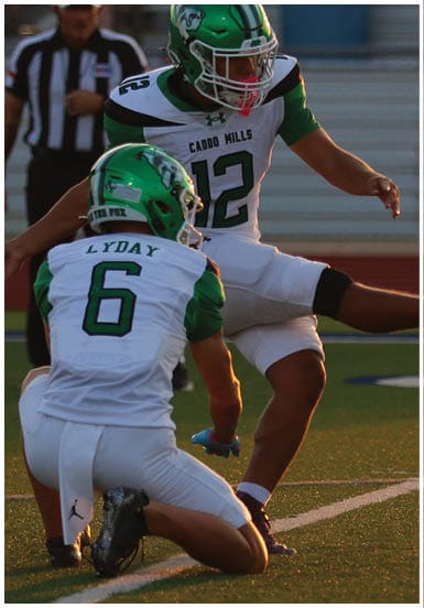 Two people in football uniforms with green helmets are on a field, with one person holding the ball and the other kicking it. Descripci n generada con IA