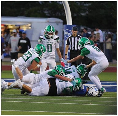 Several people in green and white football uniforms are tackling a person in a blue and white uniform on a football field during a game. Descripci n generada con IA