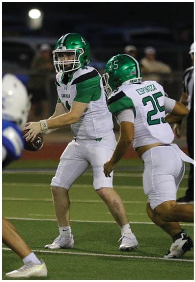 A person in a green and white football uniform is holding a football and preparing to pass while another person in a similar uniform stands nearby and an opposing player in a blue uniform approaches. Descripci n generada con IA
