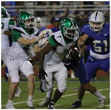 A person in a white and green football uniform is running with the ball while two people in blue football uniforms attempt to block or tackle on a football field with spectators in the background. Descripci n generada con IA