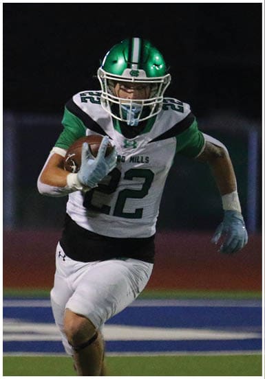 A person wearing a green and white football uniform and helmet is holding a football while running on a field at night. Descripci n generada con IA