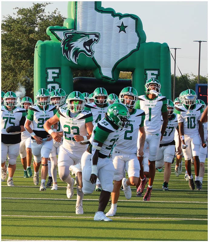 A group of people in white and green football uniforms are running onto a field from an inflatable tunnel shaped like the state of Texas with a wildcat logo. Descripci n generada con IA
