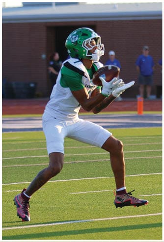 A person wearing a green and white football uniform is catching a football on a field with a brick wall and three people standing in the background. Descripci n generada con IA