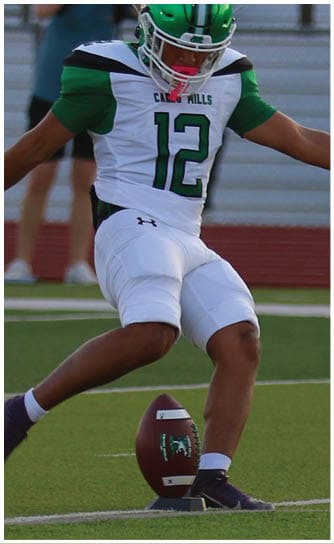 A person wearing white athletic pants and dark cleats is preparing to kick a football placed on a tee on a green field. Descripci n generada con IA