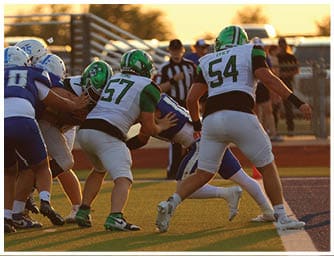 People wearing football uniforms are engaged in a close physical play on a grassy field during a game. Descripci n generada con IA