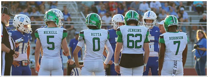Four people in white sports uniforms with green helmets stand on a field facing away from the camera while others and spectators are visible in the background. Descripci n generada con IA