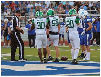 Two people in white and green football uniforms with helmets stand on a field near a referee wearing a black and white striped shirt. Descripci n generada con IA