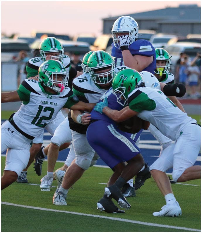 People in football uniforms are engaged in a tackle on a field with a person in casual clothing taking photos in the background. Descripci n generada con IA