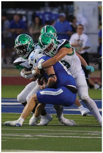 Two people in green and white football uniforms tackle a person in a blue uniform holding a football on a field. Descripci n generada con IA