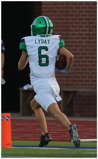 A person wearing a green and white football uniform with the name Lyday and number 6 is running with a football toward an orange pylon on a sports field while a person in a dark uniform stands in the background near a brick wall. Descripci n generada con IA