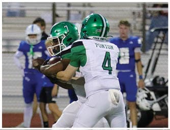 Two people in green and white football uniforms are engaged in a tackle during a game with referees and other players in the background. Descripci n generada con IA