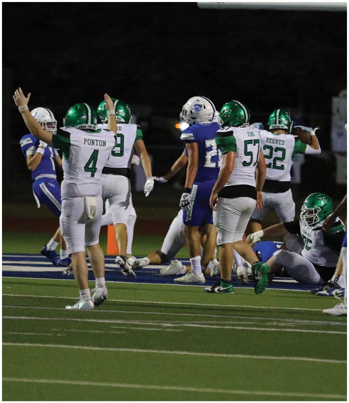 People in football uniforms are on a field with some standing and one sitting on the ground during a nighttime game. Descripci n generada con IA