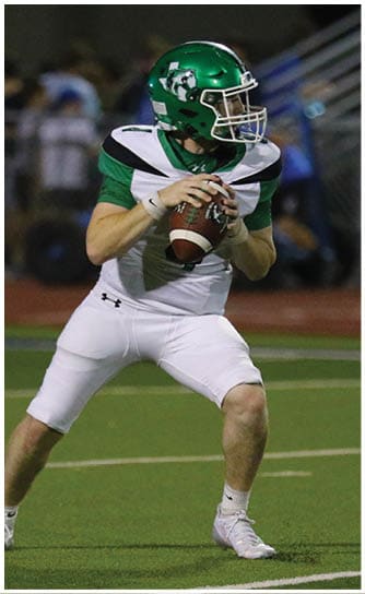 A person wearing a green helmet and white football uniform is holding a football and preparing to throw it on a field. Descripci n generada con IA