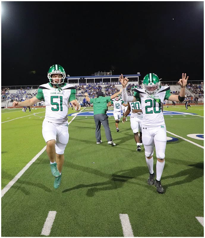 A person wearing a green and white football uniform with the number 51 is jumping on a football field at night with other players and a coach in the background. Descripci n generada con IA