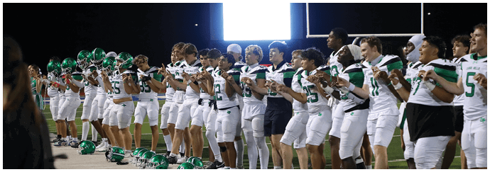 A group of people in white sports uniforms with green helmets stand in a line on a field at night, clapping with a scoreboard visible in the background. Descripci n generada con IA