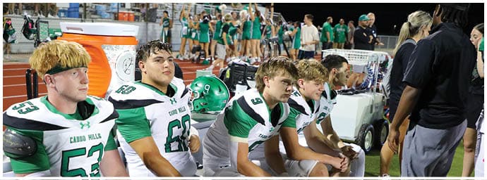 People in football uniforms sit on a bench on a sports field while another person stands nearby. Descripci n generada con IA