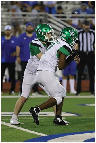 Two football players in green helmets and white uniforms are on a field with one player handing off the ball to the other while people and a referee stand in the background. Descripci n generada con IA