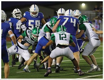 Two people wearing green and white football uniforms and helmets are positioned closely together, appearing to be engaged in a play on the field. Descripci n generada con IA