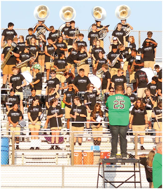 A group of people wearing black shirts with “Caddo Mills" printed on them are playing various musical instruments in bleachers while a person in a green shirt with the name "Ryan" and number 25 stands on a platform conducting them. Descripci n generada con IA