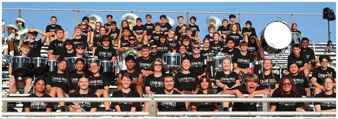 A large group of people wearing matching black shirts are seated in bleachers with many holding musical instruments such as drums and brass instruments, suggesting a band or musical group gathering outdoors. Descripci n generada con IA