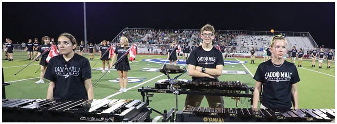 Three people wearing matching black shirts stand behind percussion instruments on a football field at night with a crowd and other performers in the background. Descripci n generada con IA