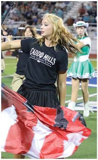 A person with long hair wearing a black shirt and skirt is holding a red flag on a sports field while another person in a green and white cheerleading outfit stands in the background. Descripci n generada con IA
