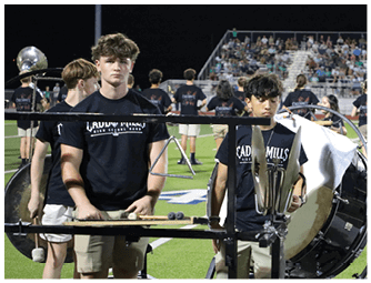 A person wearing a black shirt plays a large drum on a sports field while other people in matching black shirts and a person in a green and white costume stand nearby with a crowd watching from bleachers in the background. Descripci n generada con IA