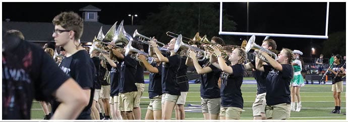 A group of people wearing matching black shirts and khaki shorts are playing brass instruments on a football field at night with a goalpost and stadium lights in the background. Descripci n generada con IA
